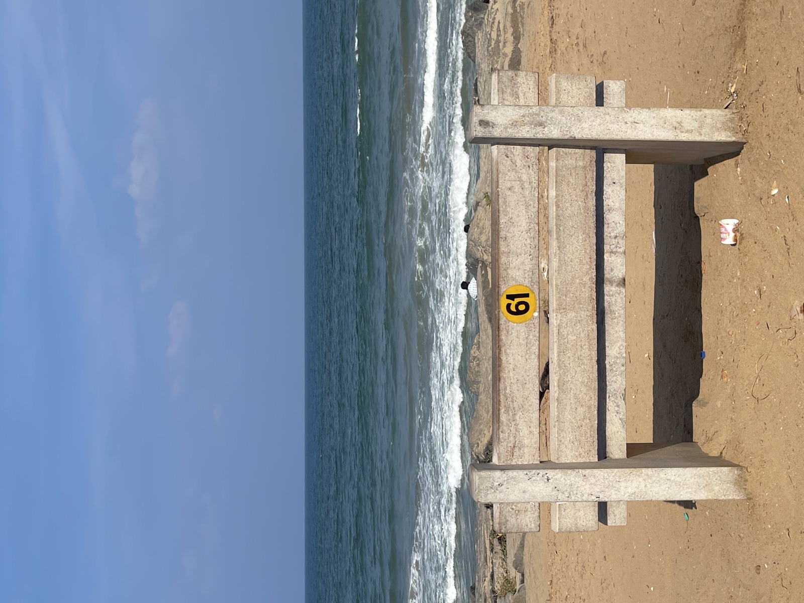 Stone bench numbered 61 facing breaking waves on a sandy beach