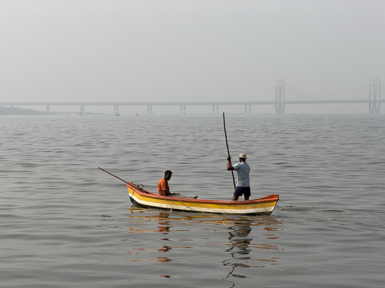 Two fishermen poling a striped wooden boat, Mumbai's Sea Link bridge hazy behind