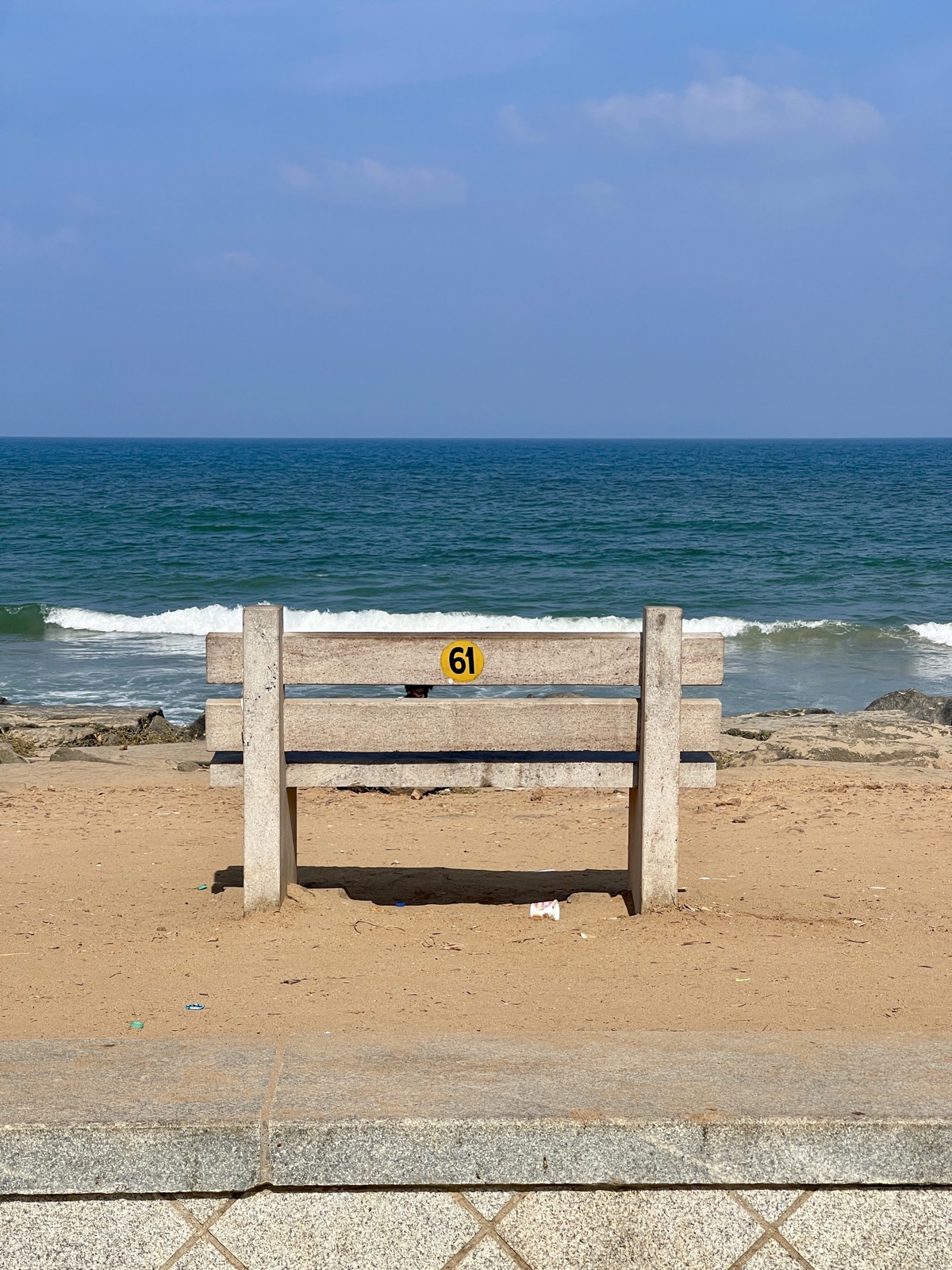 Beachside stone bench numbered 61 centered before a wave breaking on empty sand