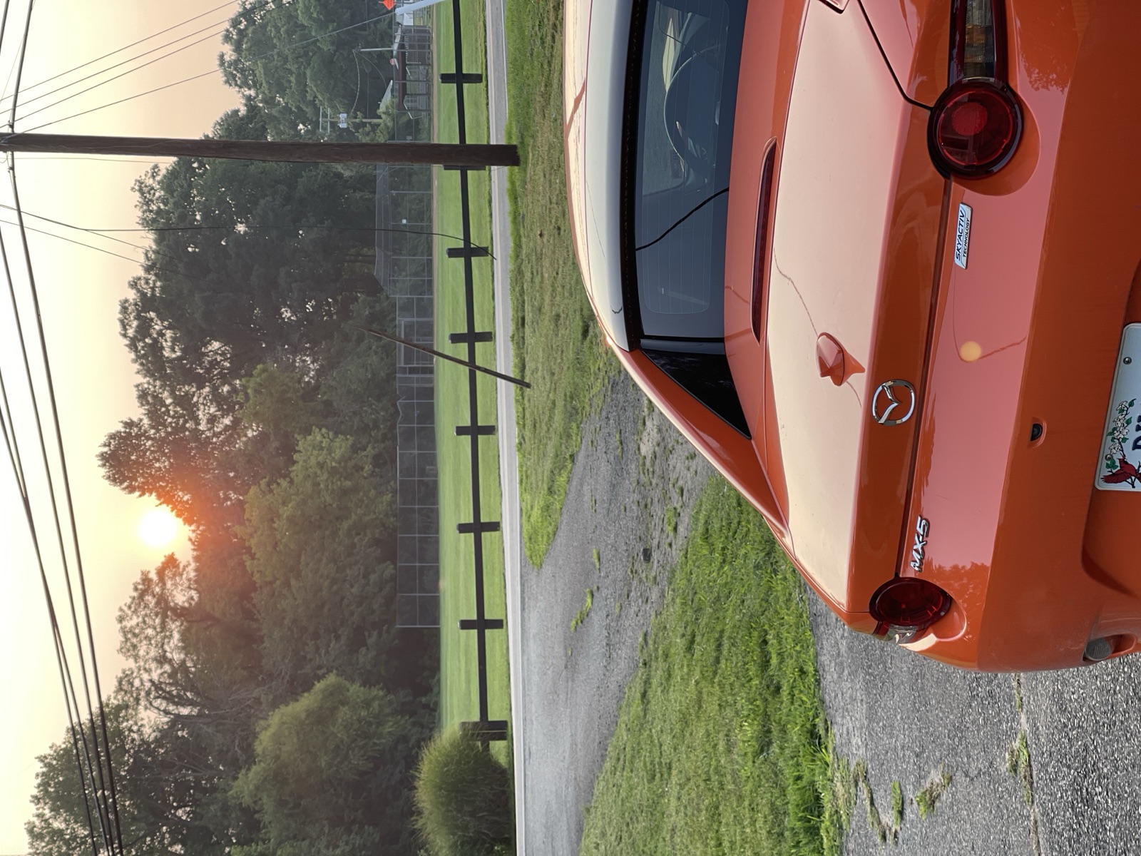 Rear of an orange Mazda MX-5 on a gravel drive as a hazy sun sets
