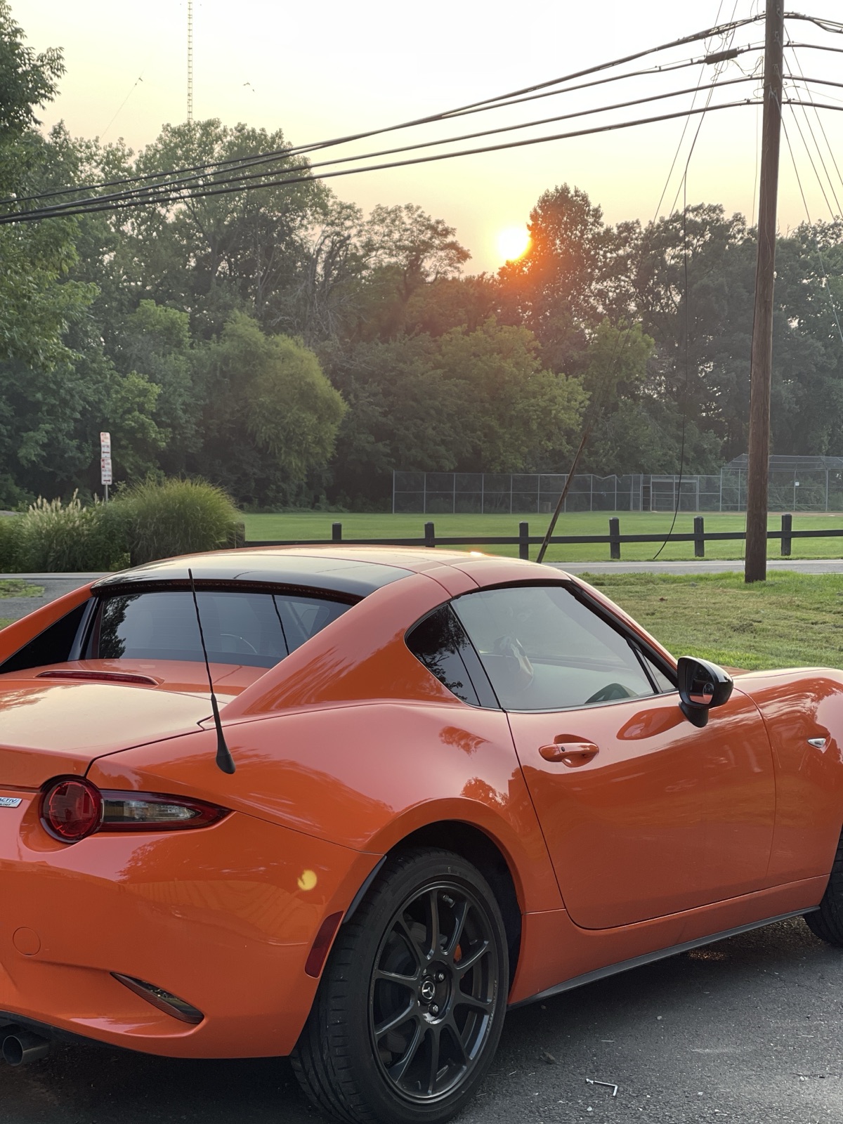 Orange Mazda MX-5 RF parked by a ballfield, sun setting through trees behind