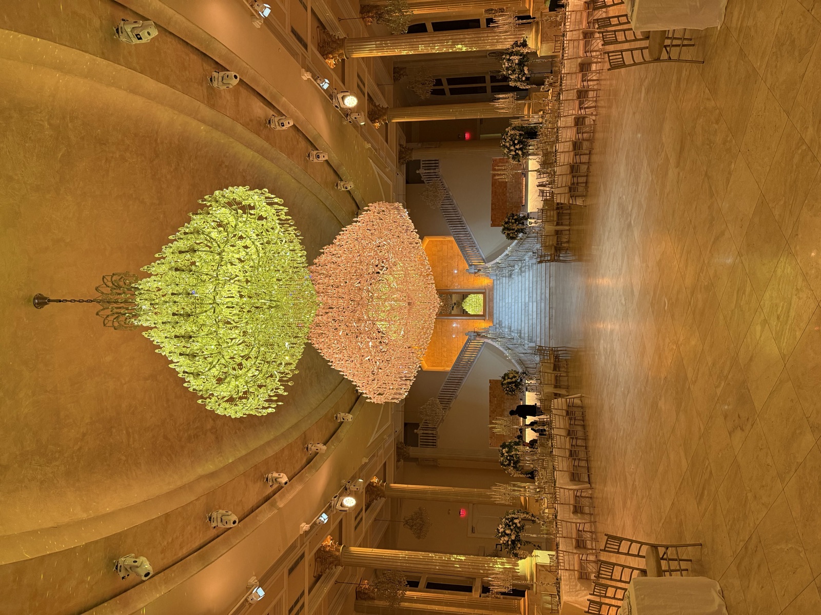 Two crystal chandeliers over a marble banquet hall set with gold chairs and flowers