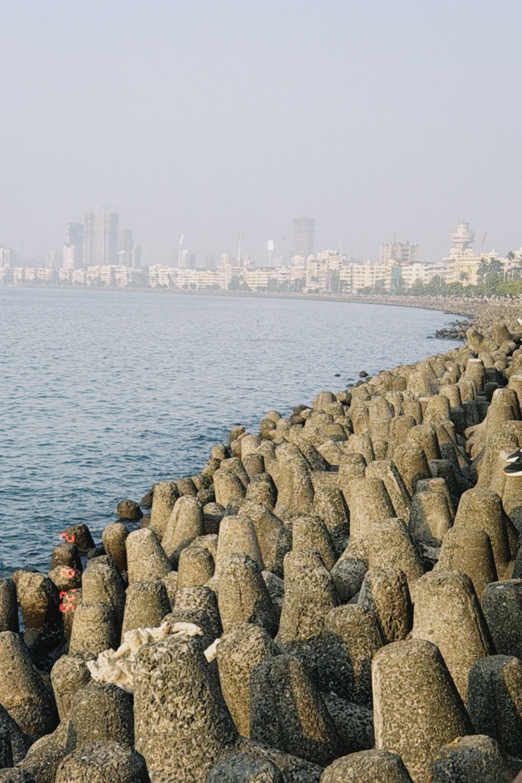 Tetrapod sea wall curving along Marine Drive toward Mumbai's hazy skyline, film grain look